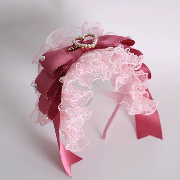 Pink lace headwear with ribbon bows, pearl accents and a heart-shaped embellishment displayed on a stand.