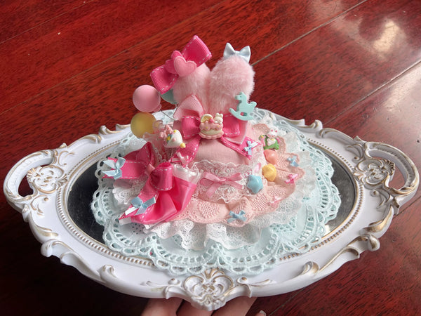 Pastel decorated mini hat with layered lace, ribbons, plush figures and round ornaments held in a hand.