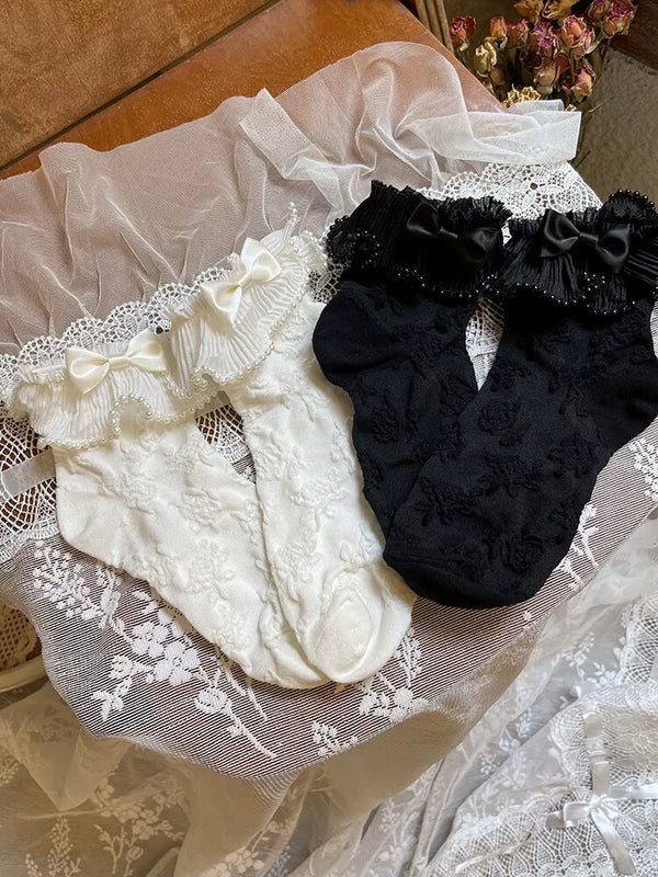 White and black short socks with lace ruffle cuffs, bows and pearl-chain trim arranged on lace fabric.