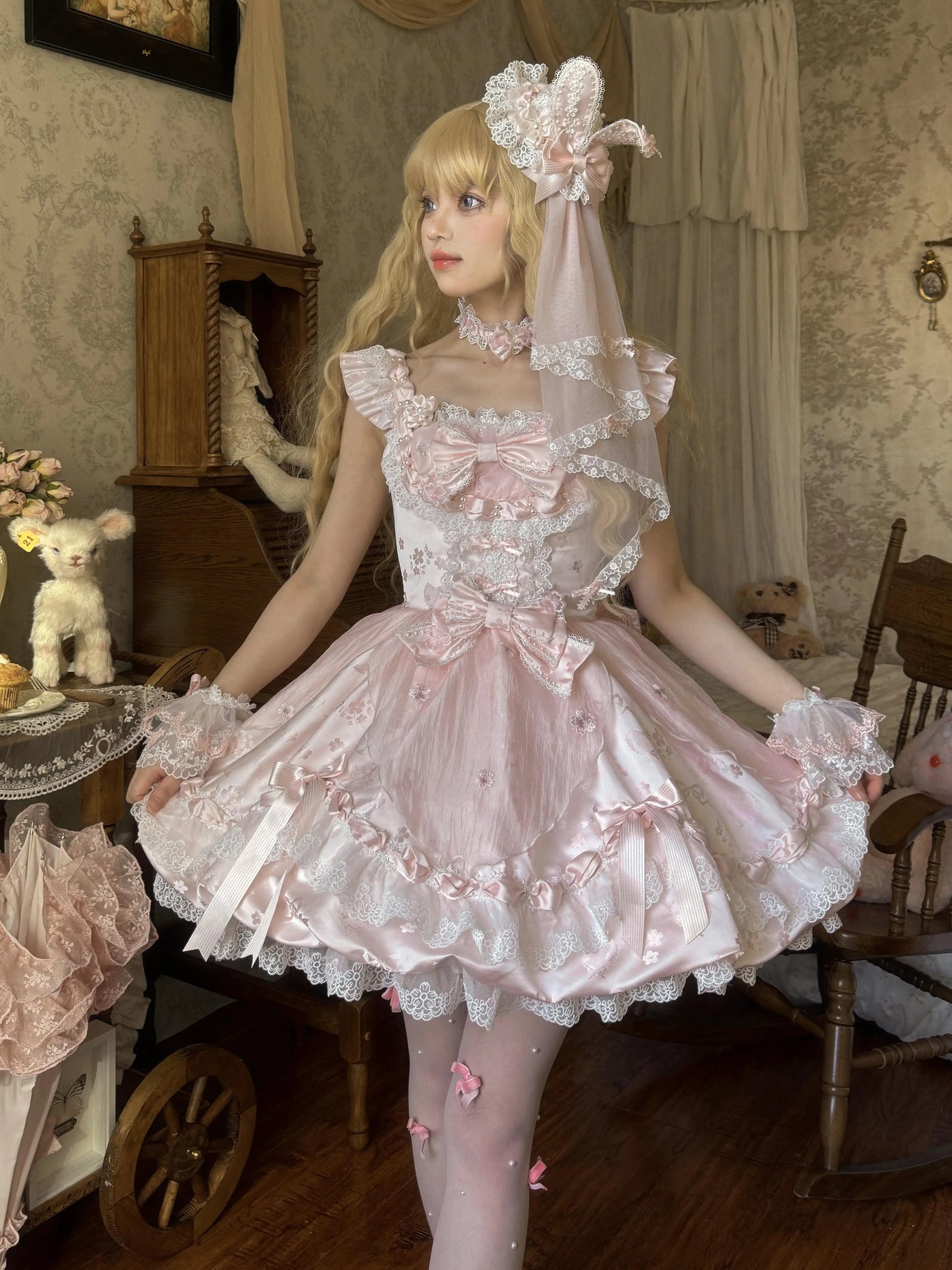 Pink JSK dress with cherry blossom motifs, layered lace and ribbon bows laid across a wooden chair.