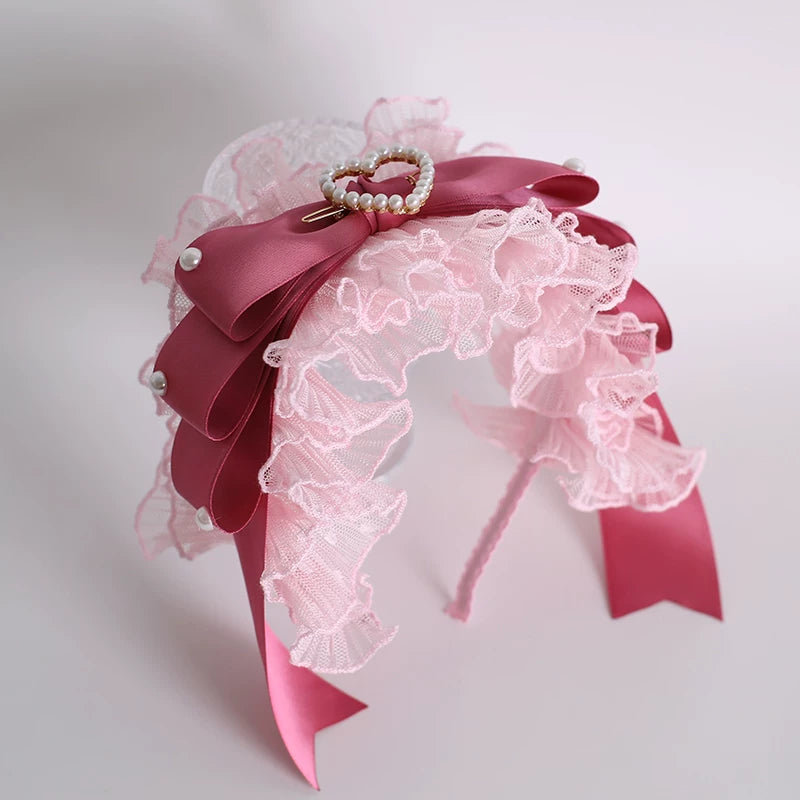 Pink lace headwear with ribbon bows, pearl accents and a heart-shaped embellishment displayed on a stand.