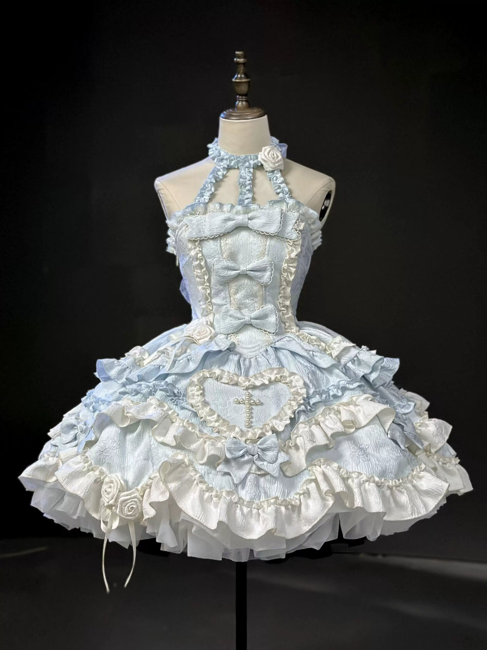 Blue-and-white JSK dress displayed on a dress form with layered bows, ruffles, roses, and a pearl cross.