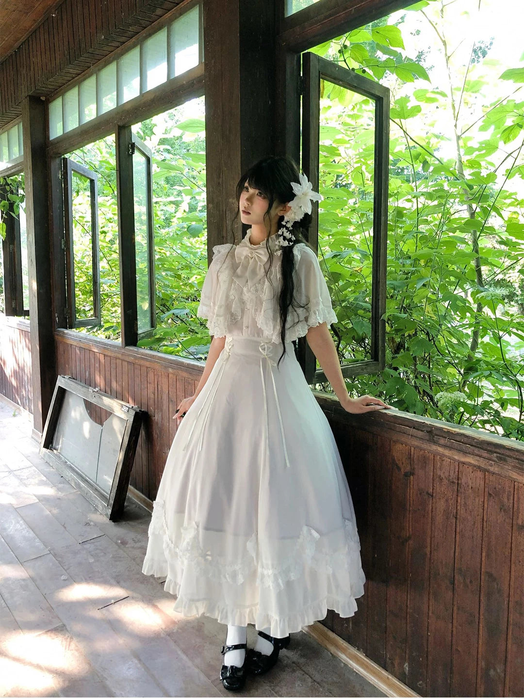 White lace Lolita dress with cape overlay worn in a leafy wooden conservatory
