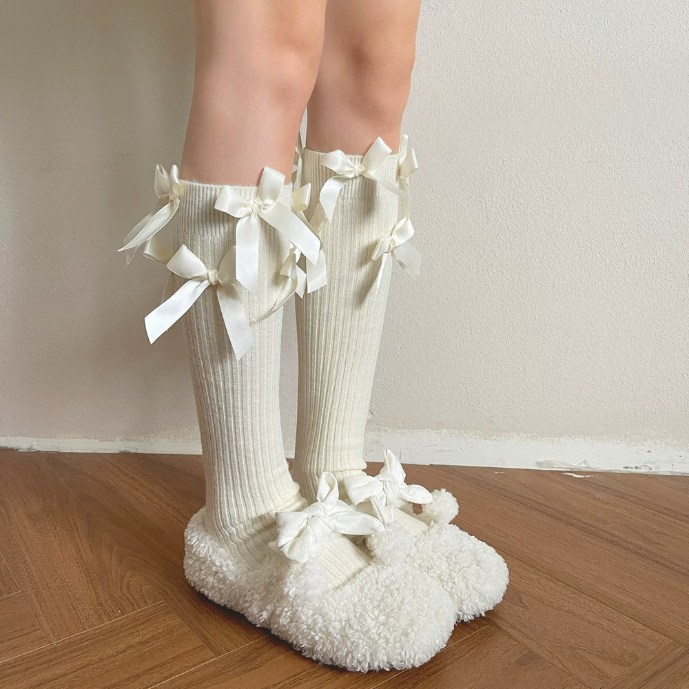 Ribbed cashmere mid-calf socks with ribbon bows and pom-pom trim styled with white slippers.