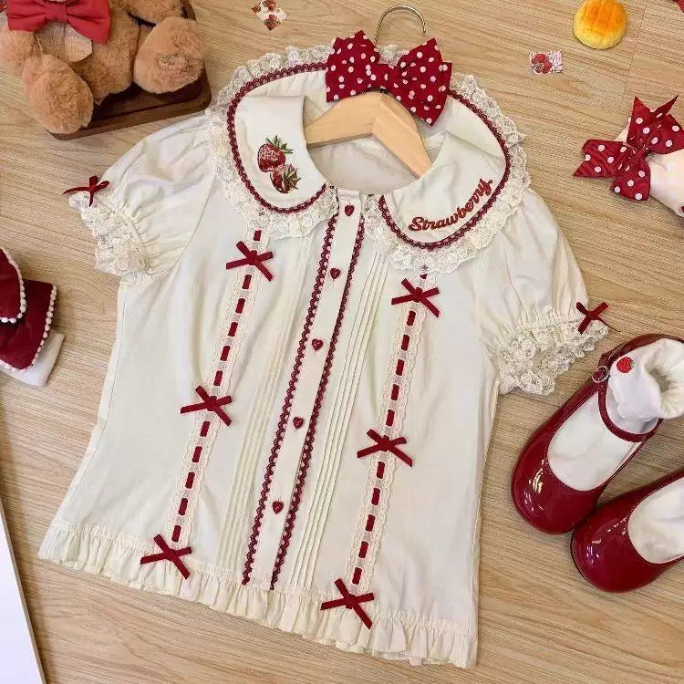 White short-sleeve blouse with strawberry embroidery, lace trim, and red ribbon accents laid flat.