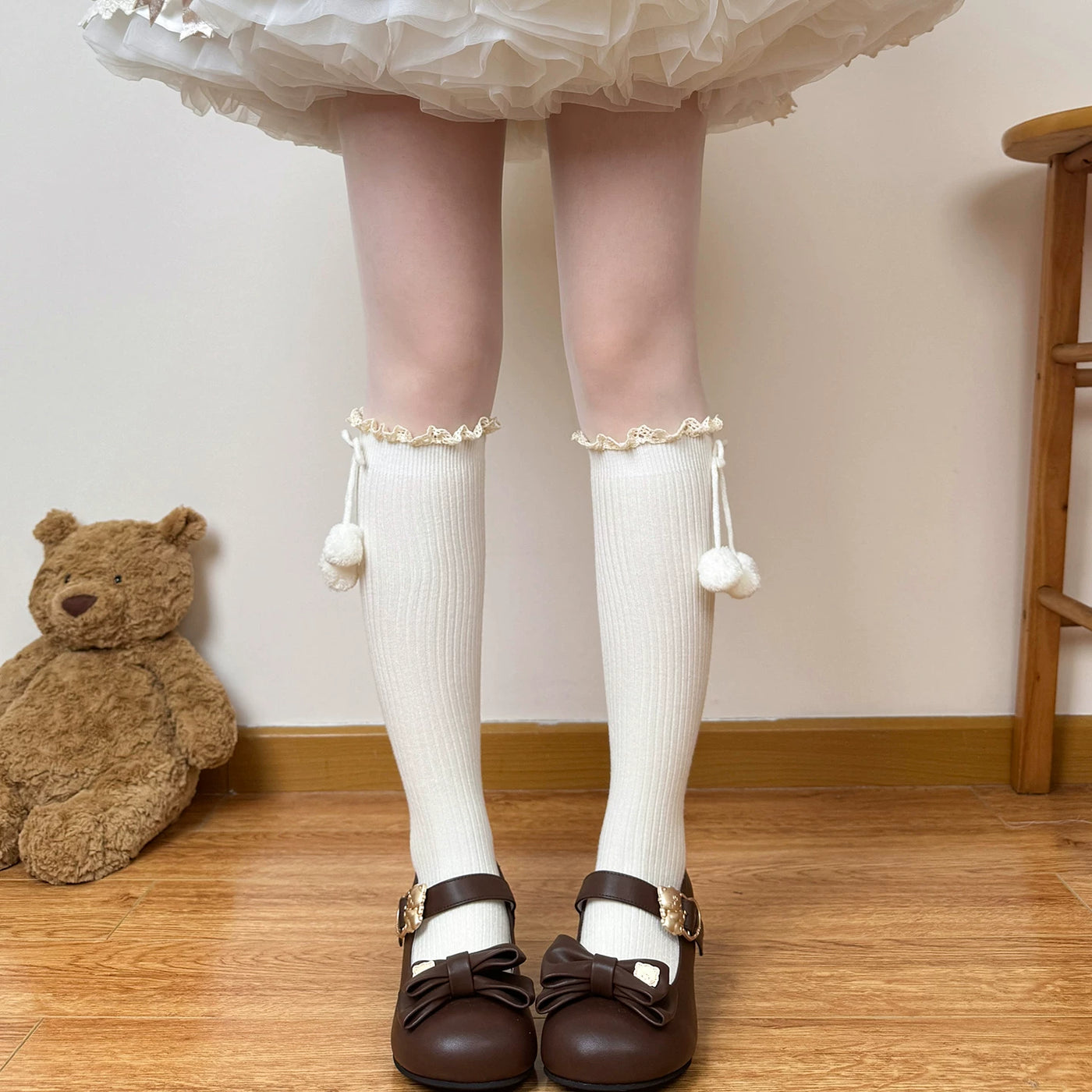 Cream-toned ribbed mid-calf scrunch socks with lace cuffs and dangling pom-poms styled with a layered Lolita skirt and brown Mary Jane shoes.