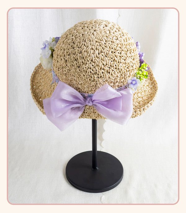 Textured woven straw hat with lavender bow and floral trim displayed on a black stand.