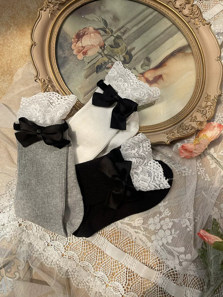 White, grey, and black ribbed mid-calf socks with lace cuffs and black bows arranged on lace fabric.