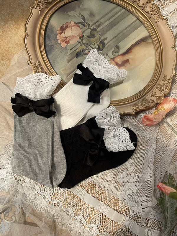 White, grey, and black ribbed mid-calf socks with lace cuffs and black bows arranged on lace fabric.