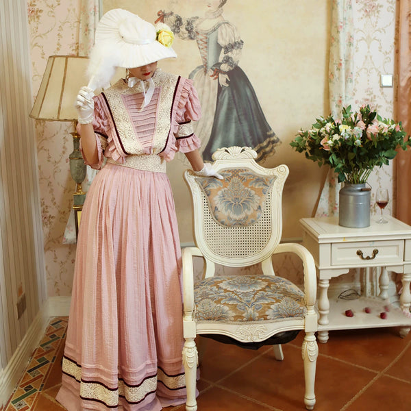 Model wearing a dust-pink Edwardian tea dress with a lace bib and full skirt.