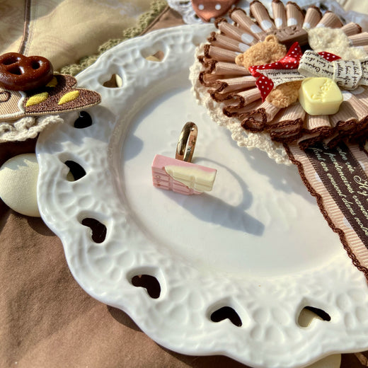 Chocolate-themed adjustable rings in assorted confection, heart, cat, teddy-bear and pastry designs displayed on a tray.