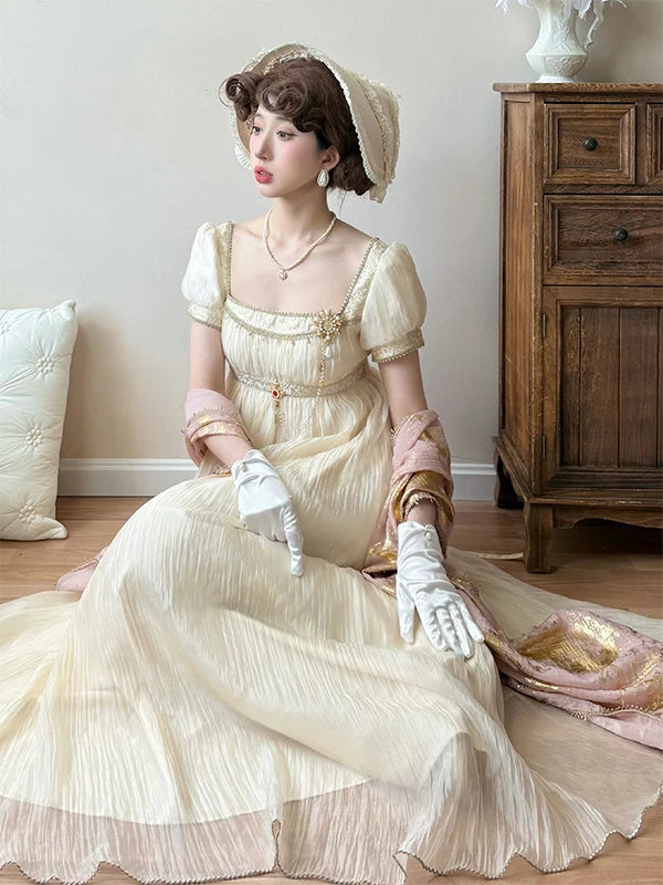 Gilded-gold empire-style dress with a square neckline, puff sleeves and pleated skirt shown on a seated model.