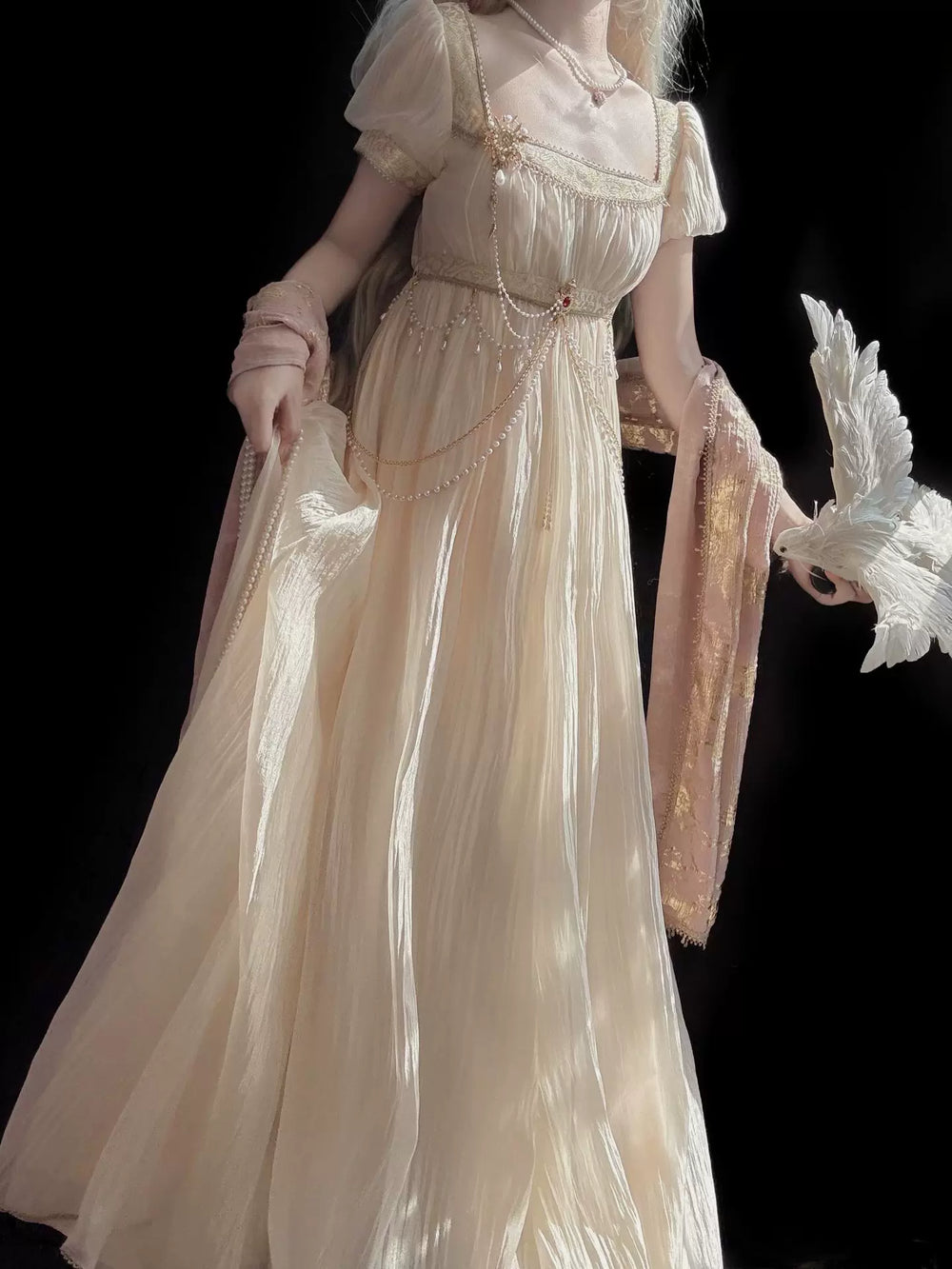 Model wearing a gilded gold-color full-length dress with puff sleeves and a draped waist chain against a black backdrop.