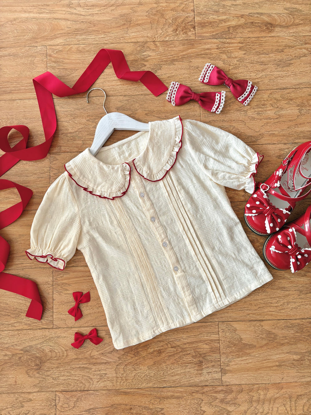 Ivory blouse with red piping, ribbon ties and lace-trimmed bows arranged on a wood floor.