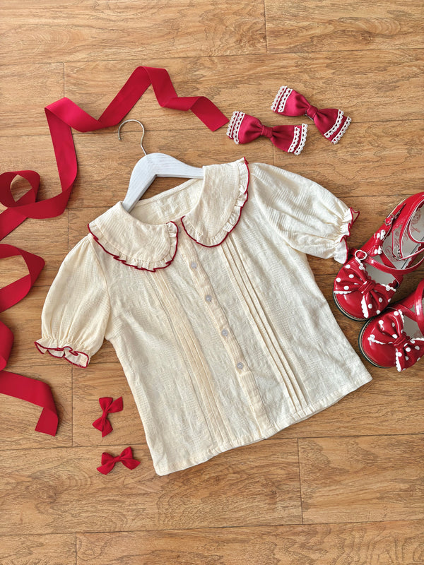 Ivory blouse with red piping, ribbon ties and lace-trimmed bows arranged on a wood floor.