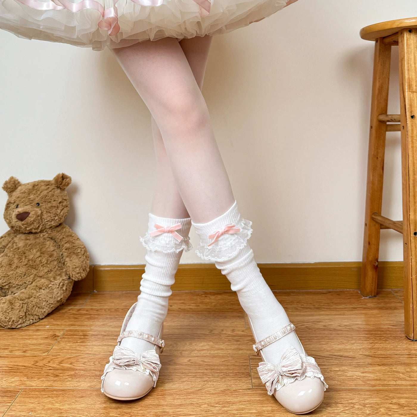 Model wearing white mid-calf slouch socks with lace ruffle cuffs and pink bows, paired with pale strap shoes.