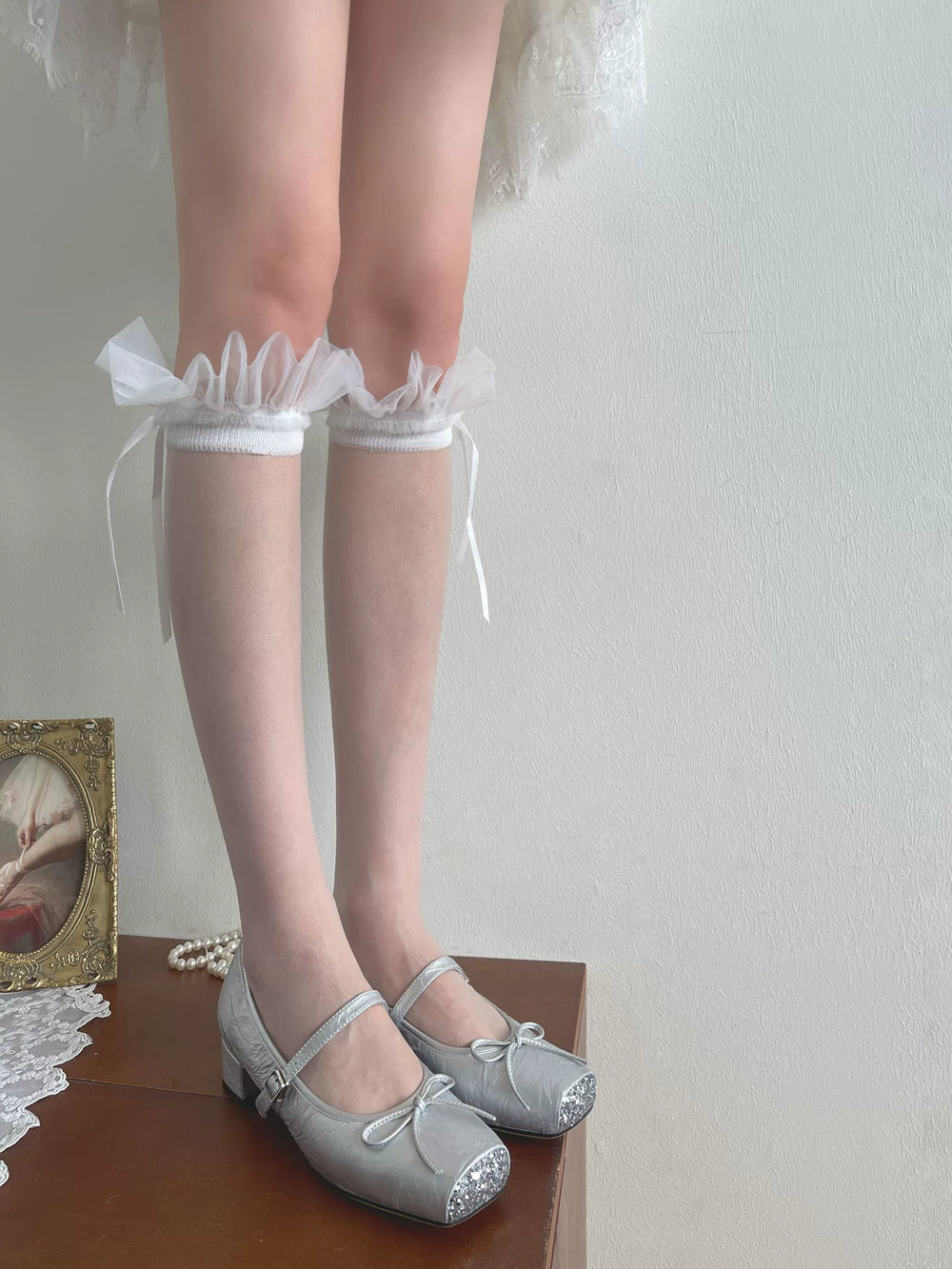 White ruffle mid-calf slouch socks shown with a pale dress hem and silver Mary Jane shoes.