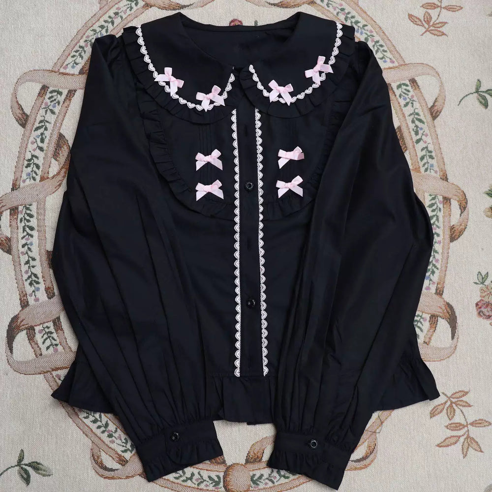 Black long-sleeve blouse with a lace-edged doll collar, ruffles and pink bows laid flat.