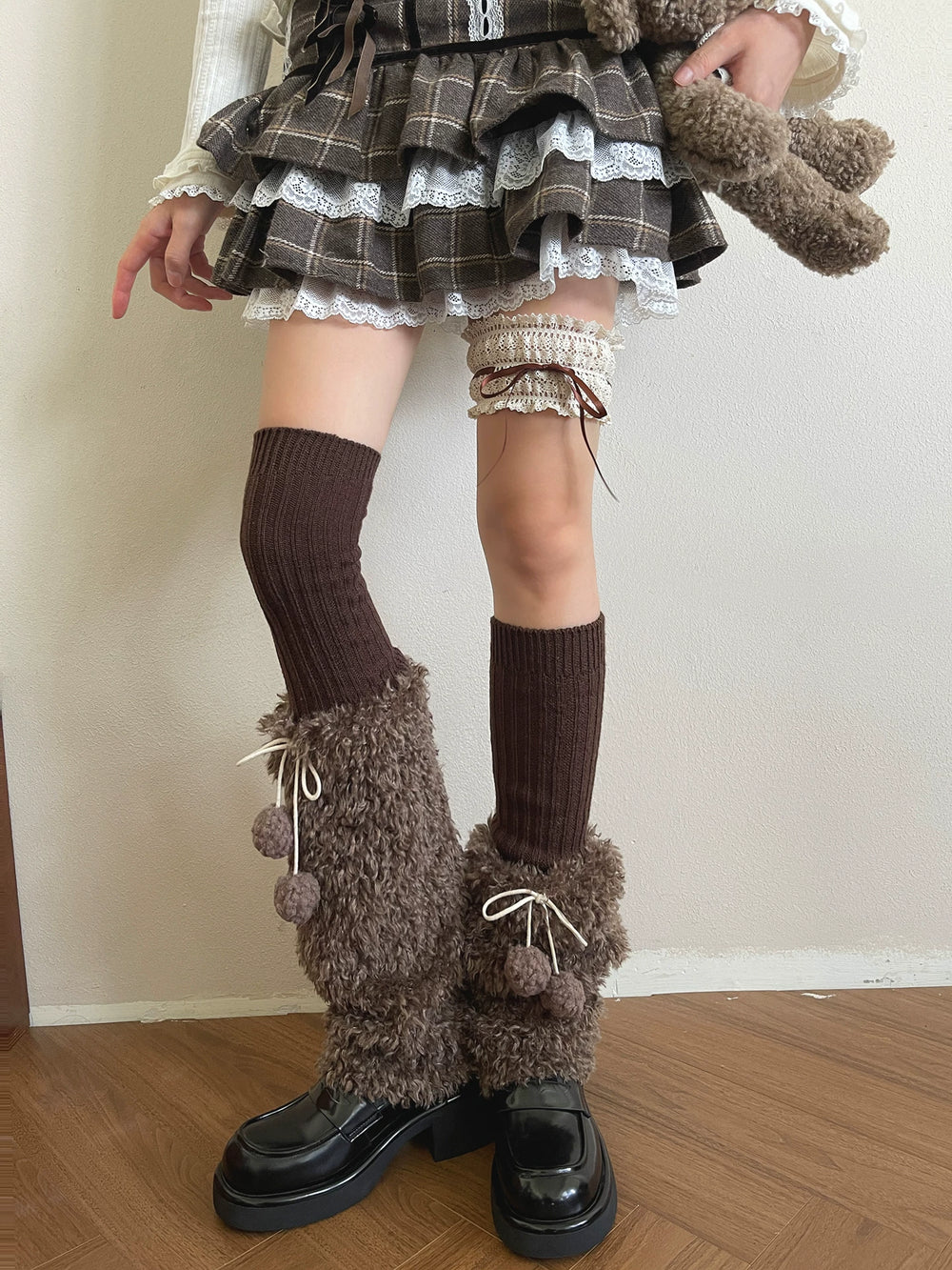 Mocha-brown plush sock covers with white ties and pom-poms styled over black shoes.