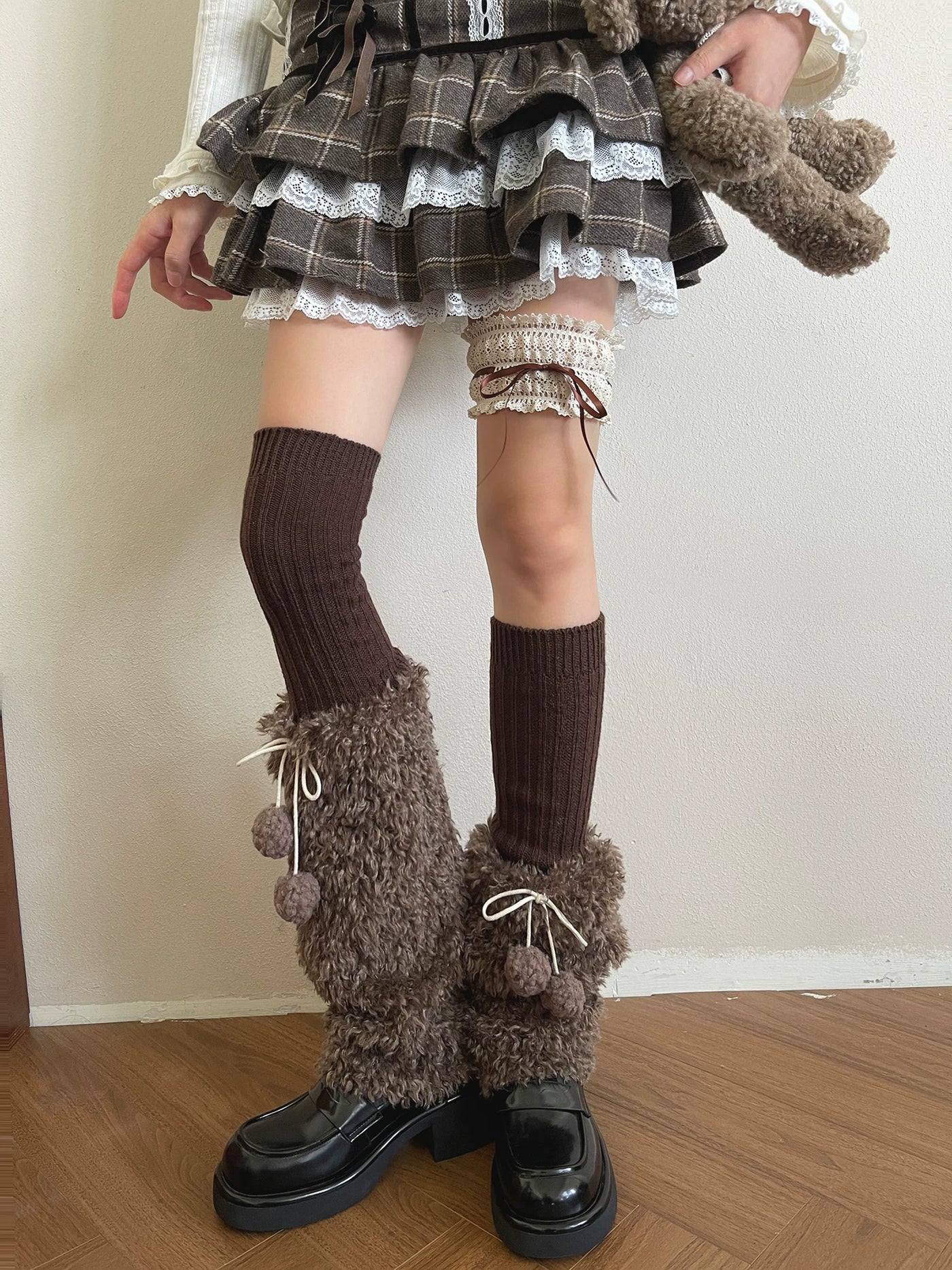 Mocha-brown plush sock covers with white ties and pom-poms styled over black shoes.
