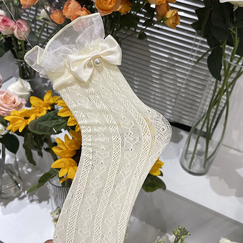 White lace mesh mid-calf sock with a ruffled cuff, satin bow and pearl detail displayed among flowers.