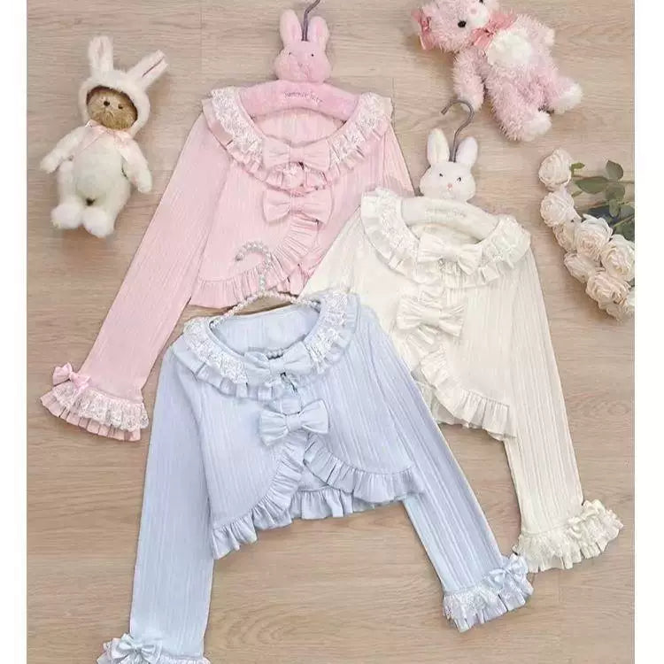 White, pink and blue ruffled knit cardigans with front bows arranged on a wood floor