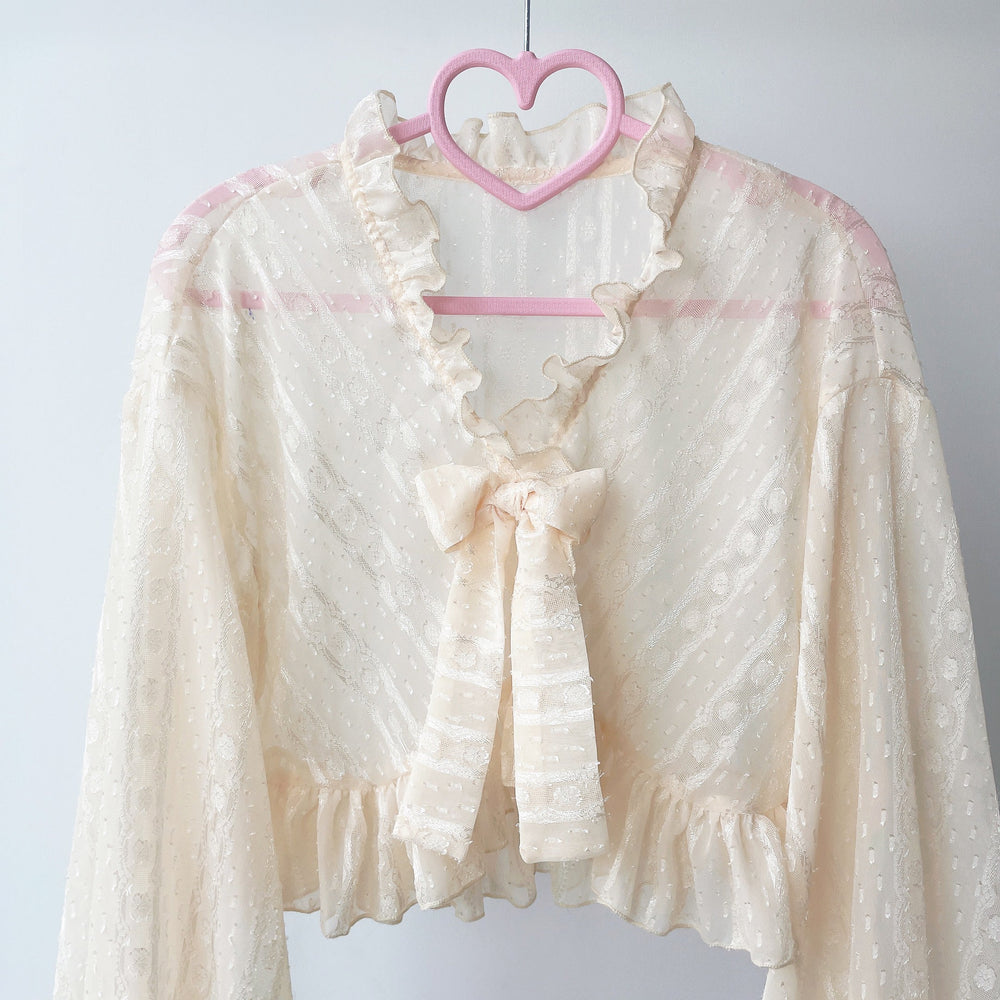 Ivory chiffon garment with a ruffled collar and bow, shown on a hanger.