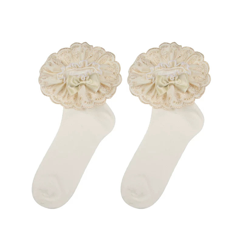 Flat-lay of cream-white short socks with champagne-gold lace edging and bow details beside a mirror.