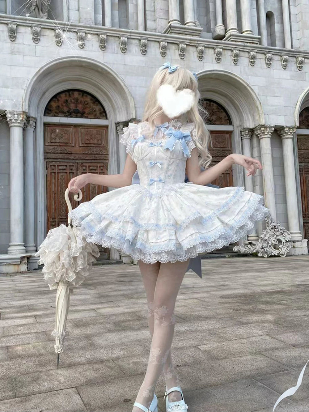 Blue-and-white layered JSK dress with rose lace and bows, shown on a model outside a grand building.