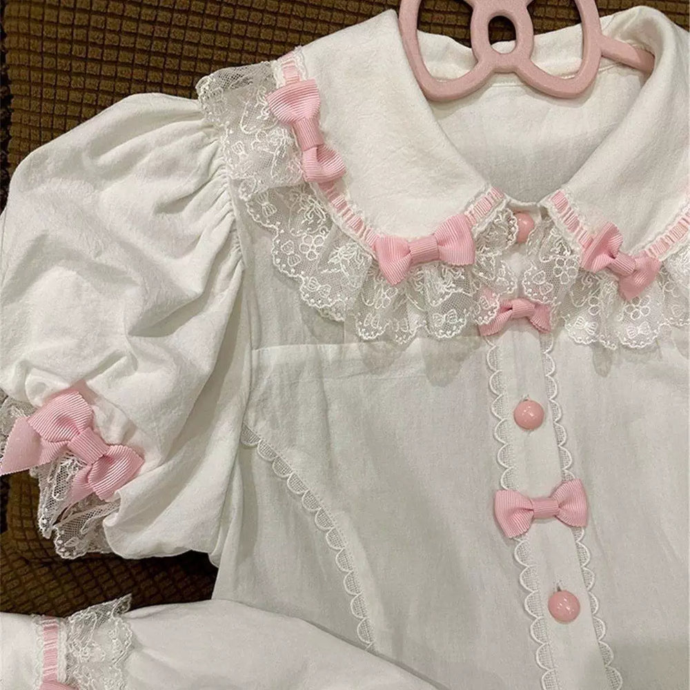 White blouse with pink bows, lace trim, puff sleeves and a ruffled hem laid flat.