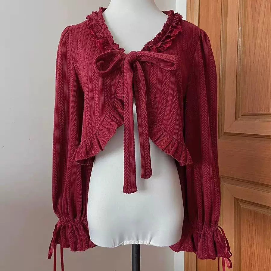 Wine-red cropped cardigan with ruffled trim, balloon sleeves and front ties laid on a wooden floor.