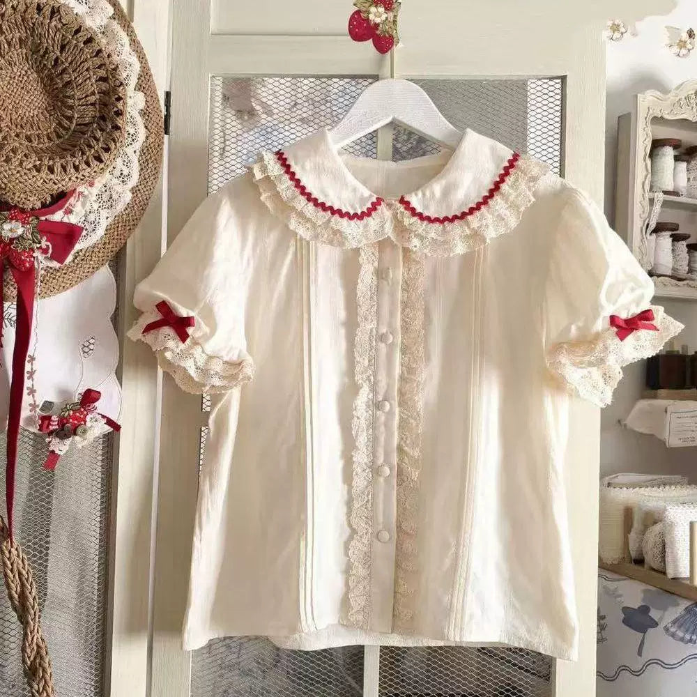 Sweetheart Dream Machine beige short-sleeve blouse with doll collar, lace trim and red bows on a hanger.