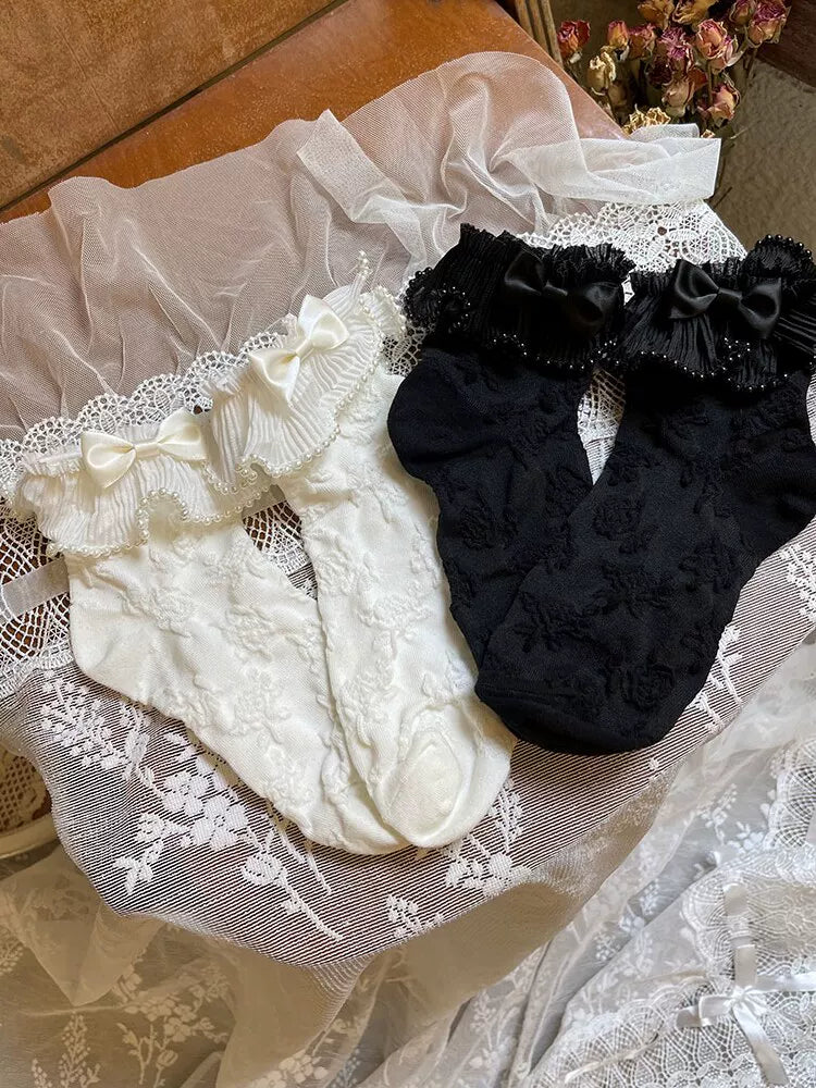 White and black short socks with lace ruffle cuffs, bows and pearl-chain trim arranged on lace fabric.
