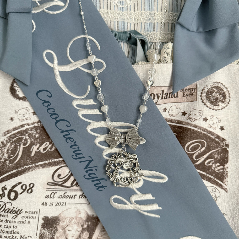 Handmade necklace with heart beads, translucent beads, a central bow and illustrated pendant on blue fabric.