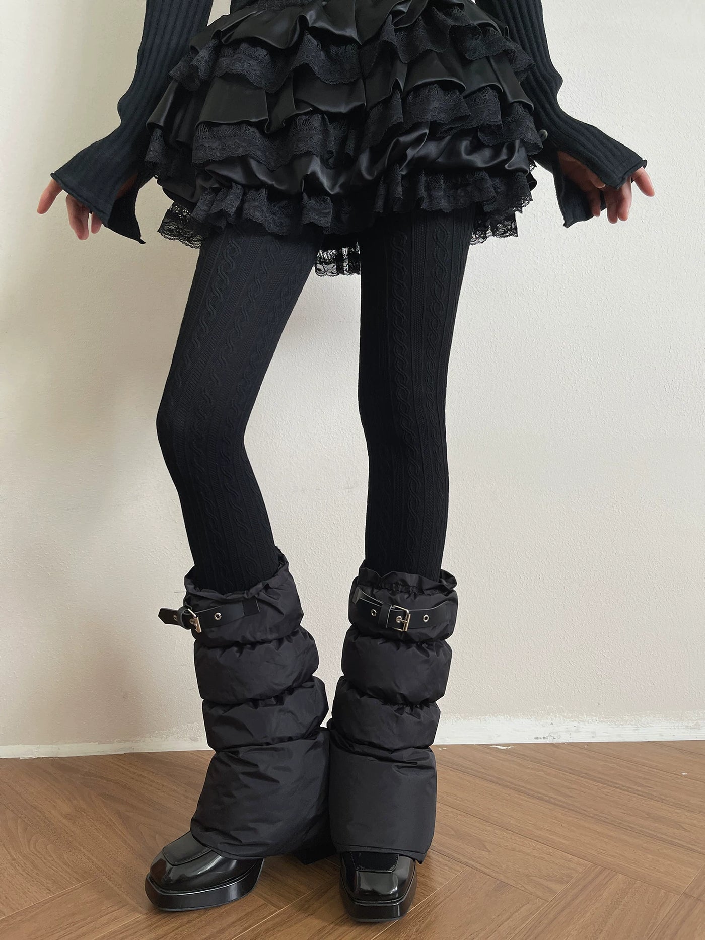 Model wearing black cable-knit tights with layered black buckle sock covers.