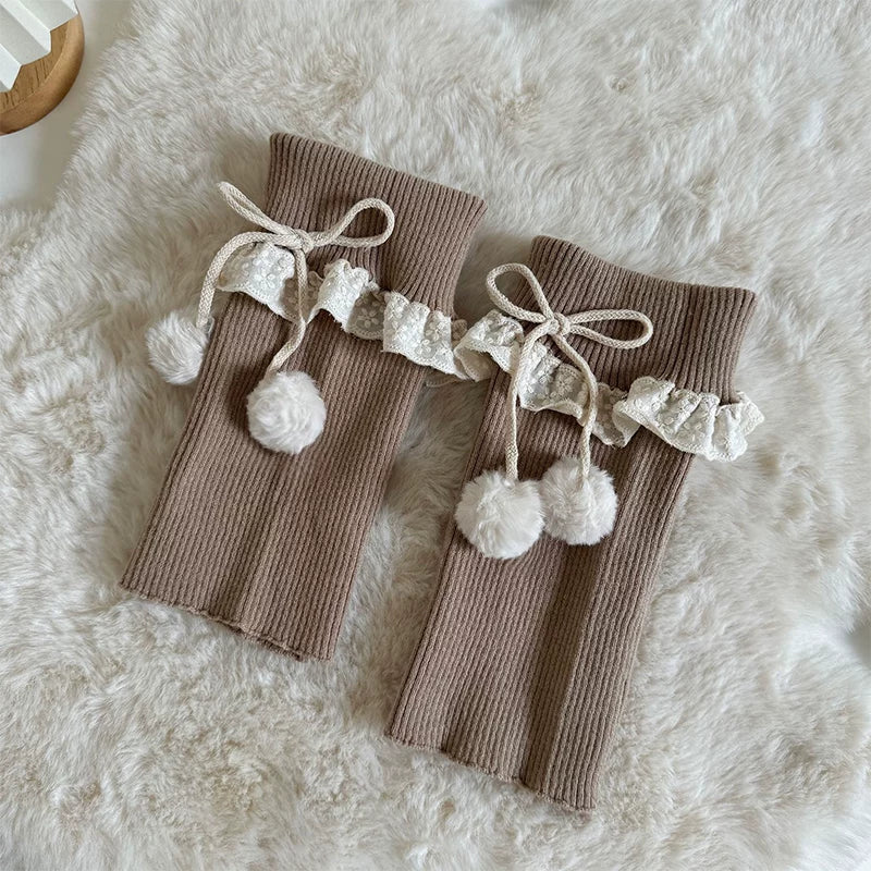 Khaki knit sock covers with lace ruffles, ribbon ties and pom-pom accents laid flat.